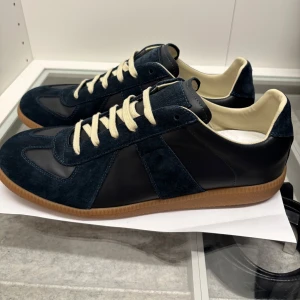 Maison Margiela blå sneakers mocka/läder - Snygga sneakers från Maison Margiela i mörkblå mocka och läder med kontrasterande vita snören och klassisk gummisula. Ikonisk design med rund tå och diskreta detaljer. Perfekta för dig som gillar stilrena och exklusiva sneakers.