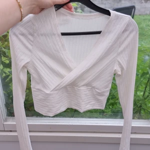 Vit topp med ribbade detaljer - Snygg croppad omlottopp i vitt med ribbade ränder och lång ärm. Toppen har en v-ringad omlottfront som ger en modern och fräsch look. Perfekt att styla med höga jeans eller kjol för en trendig outfit.