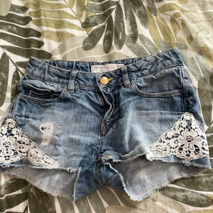 Jeansshorts med spets från Detroit - Snygga blå jeansshorts från Detroit med råa kanter och vita spetsdetaljer på sidorna. Klassisk femficksmodell med knapp och dragkedja framtill. Perfekta för sommaren och ger en cool, avslappnad vibe.