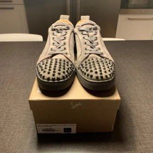 Grå mocka sneakers från Louboutin - Snygga grå sneakers i mocka från Christian Louboutin med ikoniska röda sulor och coola nitar på tån. Skorna har snörning och en rund tå, samt lyxig läderinsida. Perfekta för dig som vill sticka ut med en edgy och exklusiv stil.