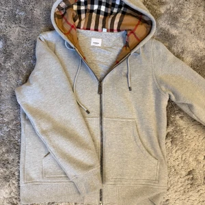 Grå Burberry Hoodie med Dragkedja - Exklusiv grå hoodie från Burberry med hel dragkedja. Endast använd en gång – i väldigt bra skick. Tidlös design, hög kvalitet och lyxig känsla i både passform och material.  🧼 Nästan som ny – fräsch & redo att användas 💬 Priset kan diskuteras – skicka gärna ett bud! 📦 Snabb leverans & smidig kontakt 