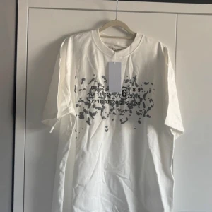 Vit t-shirt Maison Margiela med tryck - Snygg vit t-shirt från Maison Margiela med unikt grått tryck och siffror framtill. Oversized passform och klassisk rund hals. Tillverkad i mjuk bomull som känns skön mot huden. Perfekt statement-plagg för dig som gillar streetstyle.