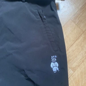 Svarta Shitkid Snowpants från Harlaut - Svarta täckbyxor från Harlaut Apparel Co. Modell Shitkid Snowpants med broderad figur på benet och diskret logga bak. Byxorna har raka ben, praktiska fickor och är fodrade för vinterbruk. Perfekta för dig som vill sticka ut i backen.