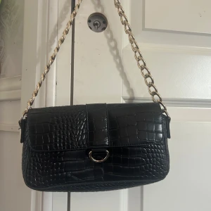 Svart axelväska med kedja från Missguided - Snygg svart axelväska från Missguided med croco-mönstrad yta och guldfärgad kedja som axelrem. Väskan har ett stilrent rektangulärt lock med metallring framtill och innerfack med dragkedja. Perfekt för dig som vill ha en trendig och elegant look.