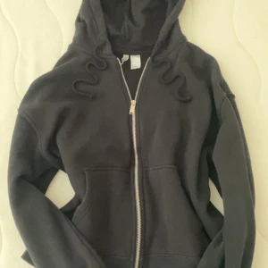 Svart hoodie med dragkedja H&M Divided - Svart hoodie från H&M Divided i storlek L. Klassisk modell med huva, snörning och hel dragkedja framtill. Mjuk insida och två fickor framtill. Perfekt för chill dagar eller när du vill ha en enkel och stilren look.