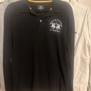 Svart långärmad pikétröja La Martina - Svart långärmad pikétröja från La Martina med klassisk krage och broderad logga på bröstet. Tröjan har gul detalj på insidan av kragen och är tillverkad i mjuk bomull. Perfekt för en stilren och sportig look.