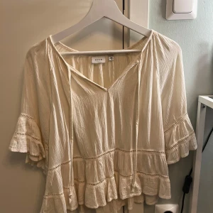 Krämvit blus med volanger från Vila - Supersöt krämvit blus från Vila med v-ringning och knytband fram. Blusen har korta volangärmar och flera volanglager nertill samt fina spetsdetaljer. Luftigt, lätt material som känns perfekt för varmare dagar.