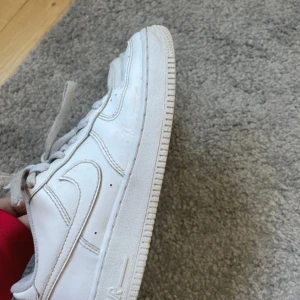 Nike Air Force 1 vita sneakers - Klassiska Nike Air Force 1 sneakers i helvitt skinn med rund tå och platt sula. Snygga detaljer som metallbricka med AF1 vid snörningen och broderad Nike Air-logga på hälen. Perfekta för dig som gillar en clean och tidlös look.