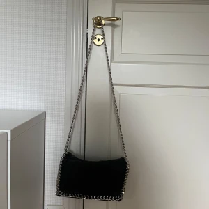 Svart axelväska med kedjedetaljer - Snygg svart axelväska med coola silverfärgade kedjor längs kanterna och som axelrem. Väskan har en mjuk, rektangulär form.Fick i present och aldrig används, hör gärna av dig för frågor💞pris kan alltid diskuteras. Nypris 499