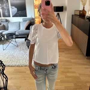 Vit blus med broderade puffärmar - Superfin vit blus med korta puffärmar i broderad bomull och rund halsringning. Ärmarna har snygga hålbroderier som ger en romantisk vibe. Perfekt till jeans eller kjol för en fräsch look.