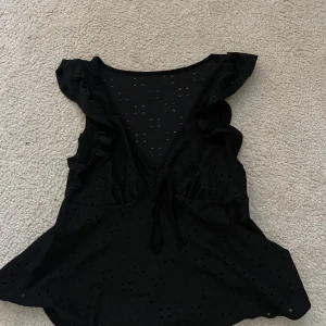 Svart blus med volang och knyt - Supersöt svart blus med volangdetaljer vid axlarna och djup v-ringning med knyt framtill. Tunt tyg med små hålbroderier ger en luftig känsla. Perfekt för dig som gillar romantiska detaljer och en feminin look.