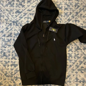 Svart hoodie från Polo Ralph Lauren - Svart hoodie från Polo Ralph Lauren med klassisk broderad logga på bröstet. Tröjan har huva med snörning, dragkedja framtill och två fickor. Tillverkad i mjuk bomull, perfekt för en chill och stilren look.Helt ny med prislapp och skriv gärna prisförslag 