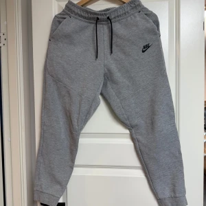 Grå Nike Tech - Snygga grå Nike Tech mjukisbyxor från Nike. Byxorna har elastisk midja med svart snörning, två sidofickor och Nike-logga på låret. Perfekta för chill eller träning, tillverkade i mjukt bomullsmaterial.