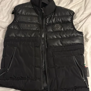 Svart dunväst från Philipp Plein - Snygg svart dunväst från Philipp Plein med quiltad design och stora logopatchar framtill. Västen har dragkedjor på fickorna och insidan är fodrad med glansigt tyg med dödskallemönster. Perfekt för dig som vill sticka ut med en exklusiv streetstyle-look.