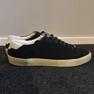 Svarta Saint Laurent sneakers i mocka - Snygga svarta sneakers från Saint Laurent i mocka med vit häl och beige sula. Klassisk låg modell med rund tå och svarta skosnören. Saint Laurent-logga på hälen ger en clean och exklusiv vibe. Perfekta för dig som gillar stilrena och lyxiga sneakers. Storlek 40 men passar 41