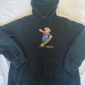 Svart Polo Bear hoodie från Ralph Lauren - Svart hoodie från Ralph Lauren med ikoniska Polo Bear-trycket på bröstet. Björnen har blå overall, röda och vita detaljer samt åker snowboard med grönt 'POLO'-tryck. Klassisk känguruficka och huva. Perfekt för dig som gillar streetstyle och sportiga vibes.