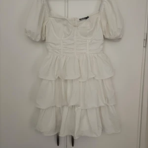 Vit volangklänning från PrettyLittleThing - Superfin vit kortklänning med volanger och puffärmar från PrettyLittleThing. Klänningen har markerad byst, rynkade detaljer och dragkedja i ryggen. Perfekt för sommarens alla tillfällen och ger en romantisk vibe.