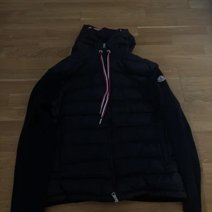 Moncler cardigan  - Svart dunjacka från Moncler med quiltad front och huva. Jackan har ribbstickade ärmar och baksida, dragkedja framtill och snygga snören i rött, vitt och blått. Moncler-logga på ärmen. Perfekt för kyliga dagar när du vill vara både varm och stilren. Storlek L passar M. Priset är inte hugget i sten så skriv till mig.