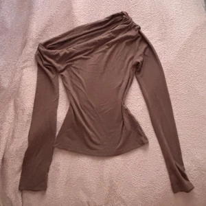 Brun offshoulder topp med långa ärmar - Säljer en brun offshoulder topp med lång ärm och figurnära passform. Tunn och stretchig, perfekt att styla med jeans eller kjol. Snyggt draperad över axlarna för en trendig look.