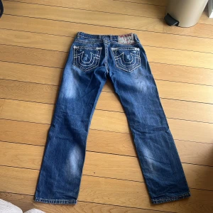 True Religion blå jeans med slitningar - Säljer ett par blå jeans från True Religion med tydliga slitningar och hål på benen. Jeansen har kontrastsömmar, klassiska bakfickor med broderad logga och patch i midjan. Raka ben och normal passform, tillverkade i bomull för en skön känsla.