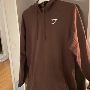 Brun hoodie från Gymshark  - Snygg brun hoodie från Gymshark i storlek M. Tröjan har huva med snörning och en stilren vit logga på bröstet. Perfekt för chill eller gymmet, med lång ärm och skön passform. Materialet känns mjukt och bekvämt mot huden. Aldrig använd