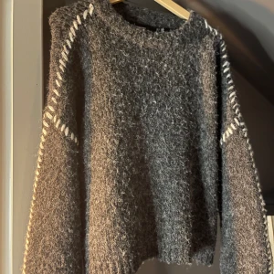 Mörkgrå stickad tröja med vita detaljer - Säljer en mörkgrå, fluffig stickad tröja med grov struktur och vita dekorativa sömmar längs axlar och ärmar. Tröjan har rund halsringning och långa ärmar, perfekt för kyliga dagar. Supermysig och trendig look!