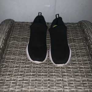 Svarta slip-on sneakers med neongul detalj - Snygga svarta slip-on sneakers från ARLINE med stickad ovandel och vit sula. Sidan har en cool neongul detalj som ger skorna en sportig vibe. Perfekta för dig som gillar stilrena men ändå unika sneakers. Lätta att ta på och av tack vare stretchig resår.