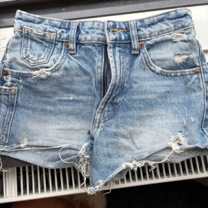 Blå slitna jeansshorts med fransar - Snygga blå jeansshorts med slitna detaljer och fransig nederkant. Klassisk femficksmodell med dragkedja och knapp. Perfekta för en avslappnad och trendig sommarlook.
