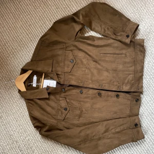 Brun mockajacka från Ljung  - Snygg brun mockajacka i veganskt material från Ljung by Marcus Larsson. Klassisk jeansjacke-modell med knappar framtill, bröstfickor med lock och krage. Perfekt för dig som vill ha en stilren och trendig look. Nypris 2499