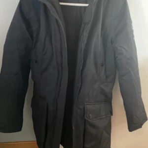 Svart parkas från Peak Performance - Säljer en svart parkas från Peak Performance med klassisk lång modell och praktiska fickor framtill. Jackan har dragkedja och är perfekt för kyliga dagar. Snygg och stilren design som funkar till det mesta. Materialet är slitstarkt och håller värmen bra.