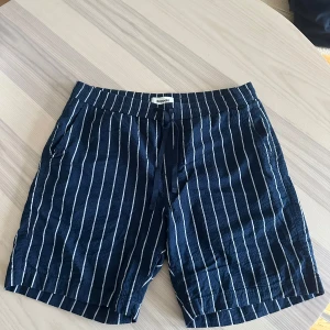 Randiga shorts från Kappahl - Mörkblå shorts från Kappahl med vita vertikala ränder. De har elastisk midja med snörning och två sidofickor samt en bakficka. Sköna och luftiga, perfekta för varma dagar. Tillverkade i mjukt bomullstyg för en avslappnad vibe. Aldrig anväda