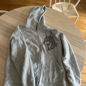 Grå hoodie med hundmotiv LT Clothing - Grå hoodie från LT Clothing med dragkedja och huva. På bröstet finns ett stort hundmotiv i svarta och silvriga nitar som ger en cool vibe. Tröjan har långa ärmar och två fickor framtill. Perfekt för dig som gillar streetstyle och vill sticka ut.
