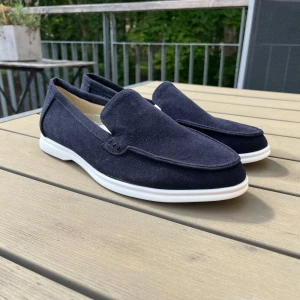 Mörkblå mockaloafers med vit sula - Stilrena loafers i mörkblå mocka med vit platt sula. Skorna har en klassisk, minimalistisk design med rund tå och är perfekta för dig som gillar en clean look. Passar till både jeans och chinos för en avslappnad men snygg stil.