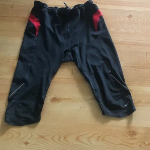 Nike FIT svarta träningsbyxor L - Svarta Nike FIT träningsbyxor i trekvartsmodell med röda detaljer och reflexdetaljer vid benen. Byxorna har en smal passform och är gjorda i ett syntetmaterial som andas, perfekt för löpning eller gym. Bakficka med dragkedja för småsaker. Känns som medium.