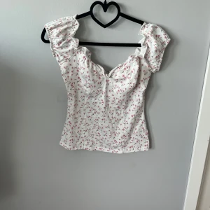 Vit blommig blus - Supersöt vit blus med små rosa blommor. Toppen har en lätt v-ringning och rynkade detaljer vid axlarna. Perfekt för varma dagar och ger en fräsch look till jeans eller kjol.