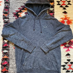 Mörkgrå hoodie från Hollister - Mysig mörkgrå hoodie från Hollister med broderad logga på bröstet. Tröjan har huva med snörning, känguruficka och långa ärmar. Perfekt för chill dagar och enkel att matcha med jeans eller jogers.