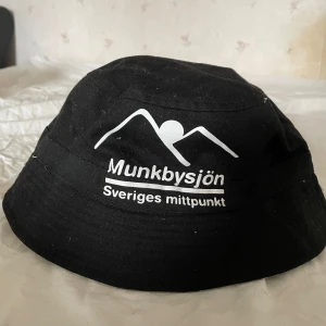 Svart bucket hat Munkbysjön - Svart bucket hat i bomull med vit tryckt logga och texten 'Munkbysjön Sveriges mittpunkt' framtill. Klassisk form med bred, nedåtlutande brätte och ljus insida. Perfekt för dig som vill sticka ut med en unik svensk touch.