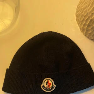 Svart Moncler mössa  - Svart mössa från Moncler i klassisk modell med uppvikt kant och broderad logotyp framtill. Tillverkad i mörkblå mjuk ull. En liten defekt på baksidan av mössan syns på andra bilden inringat i blått, men inget synligt på nån meters avstånd. Vilket gör att jag fortfarande tycker att mössan är i bra skick! Mössan är såklart äkta. 