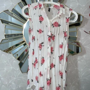 Vit blommig ärmlös skjorta  - Superfin vit skjorta från H&M med rosa blommor. Ärmlös modell med krage och lätt, flowy passform. Perfekt för varma dagar och ger en romantisk vibe.