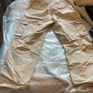 Beiga cargobyxor från FSBN, storlek M - Säljer ett par beiga cargobyxor från FSBN i storlek M. Byxorna har flera stora fickor på sidorna och bak, samt patchdetalj med CNTMP-logga. Tillverkade i bomull för en skön och avslappnad känsla. Perfekta för en chill och street-inspirerad look.