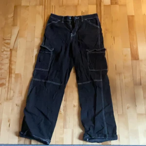 Svarta cargopants med kontrastsömmar - Svarta cargopants med bred passform och vita kontrastsömmar. Byxorna har stora fickor på sidorna och bak. Byxorna ska sitta lite baggy och brett. Välanvända och har därför ett par små hål nere vid fötterna då de suttit lågmidjat på mig men är egentligen mid-rise. 