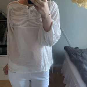Vit blus från Fik med knappar - Säljer en luftig vit blus från Fik med fina veckade detaljer och små knappar framtill. Blusen har långa ärmar med resår vid handlederna och dekorativa sömmar längs ärmarna. Perfekt för en fräsch och stilren look.