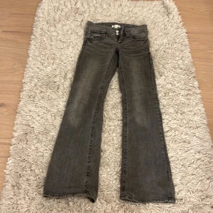 Grå bootcut jeans med dubbla knappar - Snygga grå jeans med bootcut passform och dubbla knappar i midjan. Klassisk femficksmodell med slitna detaljer och bredare benslut. Perfekta för dig som gillar en retro vibe och vill ha ett par jeans som sticker ut lite extra.