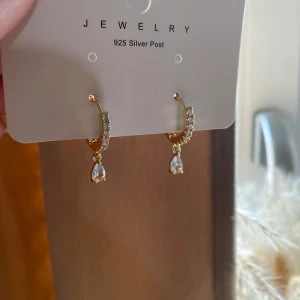 Guldplätterade örhängen  - Helt oanvända och nya guldiga örhängen med små glittriga stenar på ringen och en droppformad klar sten som hänger ned. Örhängena ger en lyxig känsla med sin eleganta design. Perfekta för att lyfta din look med lite extra bling. (Jag står inte för retur eller för postens slarv)