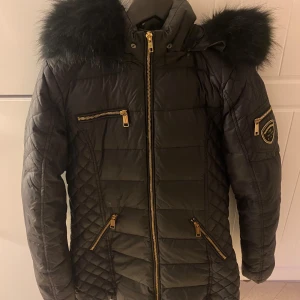 Svart Rockandblue dunjacka med fuskpäls - Snygg svart dunjacka från Rockandblue med quiltade detaljer, guldiga dragkedjor och stor huva med fluffig fuskpäls. Jackan har flera fickor med dragkedja och en patch på ärmen. Perfekt för kalla dagar när du vill vara både varm och stilren. Lite defekter så som hål i ficka och små små hål vi ärmen som syns på bild. 