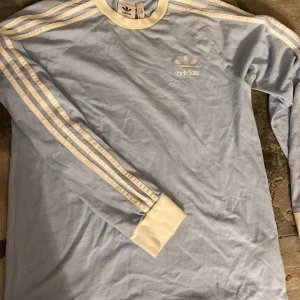 Ljusblå Adidas långärmad tröja M - Säljer en ljusblå långärmad tröja från Adidas med klassiska vita ränder längs ärmarna och vit ribbad krage. Tröjan har Adidas-logga broderad på bröstet och är gjord i mjuk bomull. 