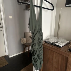 Olivgrön omlottklänning från Zara - Supersnygg olivgrön kortklänning från Zara med djup v-ringning och rynkade detaljer längs hela framsidan. Klänningen är ärmlös och har en tight passform som framhäver figuren. Perfekt för dig som vill sticka ut med en cool och trendig look.