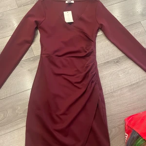 Vinröd omlottklänning från NLY One - Supersnygg vinröd omlottklänning från NLY One med lång ärm och v-ringning. Klänningen har draperad detalj i midjan och figurnära passform som framhäver siluetten. Perfekt för dig som vill sticka ut med stilren elegans.