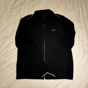 Svart zipjacka från Hugo Boss - Svart zipjacka från Hugo Boss med guldfärgad logga på bröstet. Jackan har långärmade stickade ärmar, slät framsida och dragkedja hela vägen. Snygga detaljer vid ärmslut och krage. Perfekt för dig som gillar stilren och clean look.