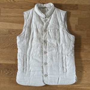 Beige quiltad dunväst från John Henric - Stilren beige dunväst från John Henric i linnemix med quiltad design. Västen har ståkrage, knäppning med fem knappar framtill och två fickor på sidorna samt en bröstficka. Perfekt för lager-på-lager och ger en clean look.